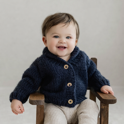 Baby wearing a navy blue knitted cardigan with brown buttons, sitting on a wooden chair against a plain background.