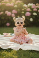 little girl wearing a Pink baby romper with cherry pattern sitting on a white blanket in the backyard