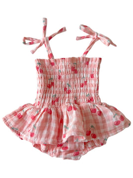 A pink and white gingham baby romper with a cherry print, featuring a smocked bodice, ruffled skirt, and tie-shoulder straps.