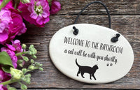 An oval white ceramic bathroom sign hanging on a rustic wooden surface, with pink flowers in the corner. The sign reads 'WELCOME TO THE BATHROOM a cat will be with you shortly.