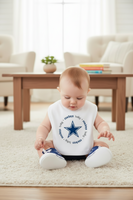 A baby sitting on a cream rug in a bright living room, wearing a white bib with a blue Dallas Cowboys star logo and the words 'Dallas baby everything'.