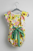 A baby girl's floral romper hanging on a black hanger. The romper features a white base with a vibrant yellow, pink, and green sunflower and floral print, accented by flutter sleeves and a large green bow at the waist.