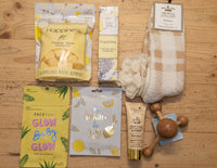 A curated wellness gift set arranged on a textured cream quilted blanket, featuring products from Faceboby, Happy Hippie, and The Pillow Standards. The set includes a yellow and green bubble bath bomb pouch, a lavender bath bomb, a beige and brown checkered towel, a wooden baby teethers massage tool, a face scrub, and a glow body scrub.