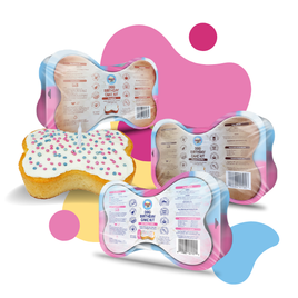 Three packaged bone-shaped cake kits for dogs are arranged on a white background with abstract pink, yellow, and blue shapes. The packages show the back labels, and one cake piece is shown with white frosting and pink and blue sprinkles.