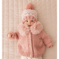 Baby wearing a pink knitted outfit with a matching hat on a beige background