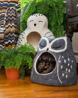 Cat peeking out from an owl-shaped pet bed with plants and a colorful blanket in the background.