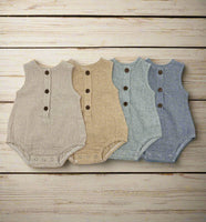 Four sleeveless baby bodysuits with three-button front closures are arranged in a row on a light wood plank background. The bodysuits are in four different colors: light beige, tan, dusty blue, and navy blue, all with a fine gingham or small-check texture.
