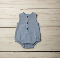 A sleeveless baby romper with a blue and white gingham pattern, featuring three brown buttons down the front, laid flat on a light wood plank background.