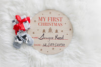 Round wooden plaque with 'My First Christmas' text, baby booties, and a small gift on a white fur background.