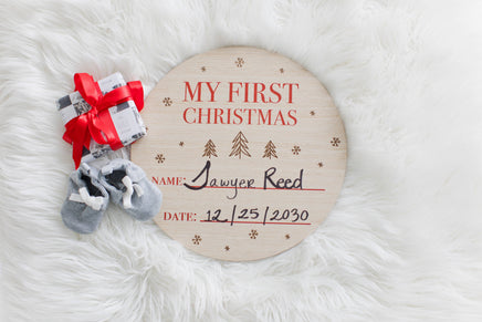 Round wooden plaque with 'My First Christmas' text, baby booties, and a small gift on a white fur background.