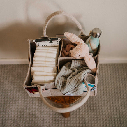 Storage diaper caddy basket with baby items including diapers, a plush toy, and a bottle on a carpeted floor.