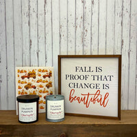Framed sign with 'Fall is proof that change is beautiful' text, two candles labeled 'Drunkin Pumpkin', and a decorative card on a wooden surface.