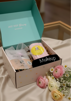 An open cosmetic gift box on draped cream satin fabric, with pink and cream flowers placed beside it. The box is light pink with small gold dots and the lid interior is mint blue, printed with the words 'JUST KEEP Glowing @KIKIKIDAY'.