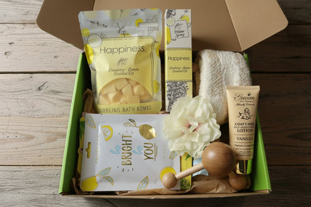 A curated self-care gift box containing a bag of Happiness Cranberry Lemon Essential Oil Bubbling Bath Bombs, a tube of Gerlin Vanillin 4 Dots Hand Lotion, a white flower, a wooden massage roller, and a facial mask, arranged in a green tray.