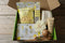 A curated self-care gift box containing a bag of Happiness Cranberry Lemon Essential Oil Bubbling Bath Bombs, a tube of Gerlin Vanillin 4 Dots Hand Lotion, a white flower, a wooden massage roller, and a facial mask, arranged in a green tray.