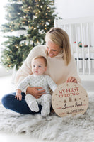 Woman holding a baby in front of a Christmas tree with a 'My First Christmas' sign.