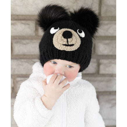 Child wearing a black knit hat with bear face design against a brick wall.
