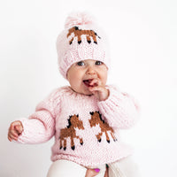 Baby wearing a pink sweater with horse designs and a matching hat on a white background