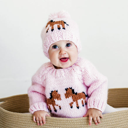 Baby wearing a pink knitted sweater and hat with horse designs on a white background