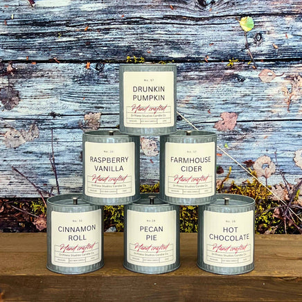 Assorted candles in tin containers with labels on a wooden surface and rustic background.
