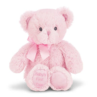 Pink teddy bear with a bow and 'Baby's First' ebroidered on his foot. with a white background