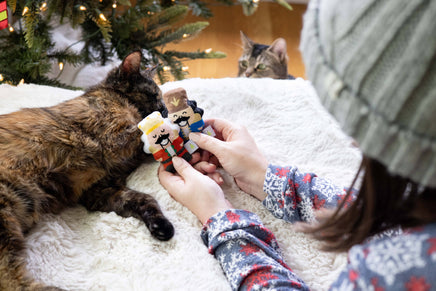 Person playing with a cat and a small figurine on a soft surface, with a Christmas tree in the background.
