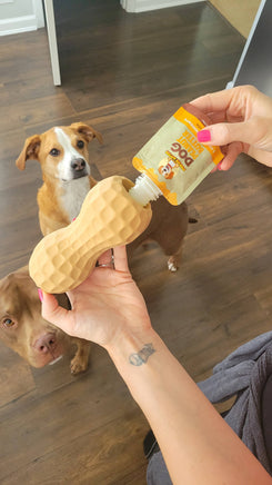 Person holding a peanut-shaped dog chew toy with a dog watching in the background.