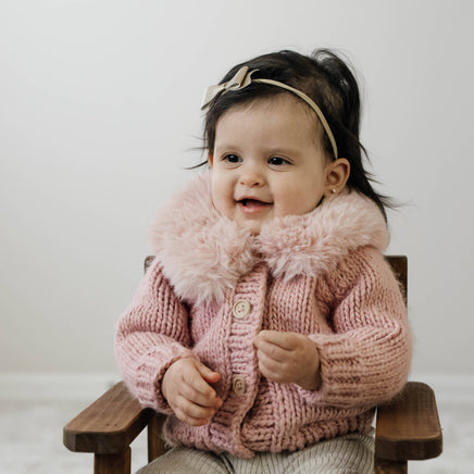 Child wearing a pink knitted jacket with fur collar sitting on a wooden chair.