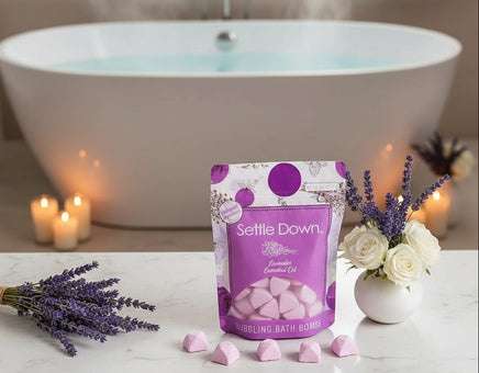 Packaging of 'Settle Down' bubbling bath bombs with lavender essential oil, surrounded by bath bombs and a vase with lavender on a light pink background.