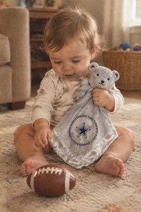 A light gray plush teddy bear security blanket featuring the Dallas Cowboys star logo and 'cowboys baby' text printed on the satin-like white blanket.
