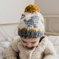Child wearing a knitted hat that has a dinosaur pattern with a pom-pom on a neutral background