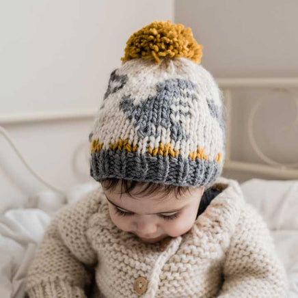 Child wearing a knitted hat that has a dinosaur pattern with a pom-pom on a neutral background
