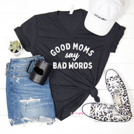 A black triblend t-shirt with the text 'GOOD MOMS SAY BAD WORDS' printed in white. The t-shirt is accompanied by blue denim jeans, a white hat, a black mug, and black and white slip-on shoes with a leopard print pattern.