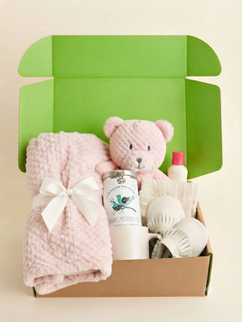 A baby gift set in an open kraft cardboard box with a bright green interior. The set includes a pink textured blanket tied with a white bow, a pink teddy bear, a white jar labeled 'Proud Parenting Products', and white baby accessories.