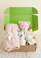 A baby gift set arranged inside an open cardboard box with a bright green interior. The set includes a light pink textured blanket tied with a white bow, a pink plush teddy bear, a white bottle labeled 'Mammoth Preservative Oil', a small white bottle with a red cap, and white mesh baby items on a light wood surface.