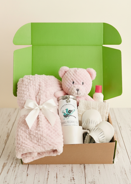 A baby gift set arranged inside an open cardboard box with a bright green interior. The set includes a light pink textured blanket tied with a white bow, a pink plush teddy bear, a white bottle labeled 'Mammoth Preservative Oil', a small white bottle with a red cap, and white mesh baby items on a light wood surface.