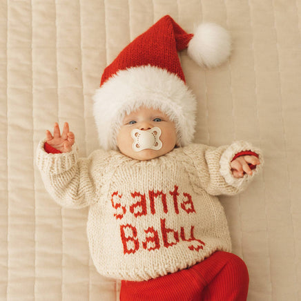 Baby wearing a 'Santa Baby' sweater and Santa hat on a beige background