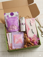 A curated gift box set displayed in an open green box on a white wood surface. The set includes a purple Seville Down bath bomb package, a white canister labeled Water Dream, a pink eyelash-themed pouch, a pink and white cloth item, two small pink sachets, a wooden rollerball massager, and a pink flower accent.