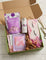 A curated gift box set displayed in an open green box on a white wood surface. The set includes a purple Seville Down bath bomb package, a white canister labeled Water Dream, a pink eyelash-themed pouch, a pink and white cloth item, two small pink sachets, a wooden rollerball massager, and a pink flower accent.