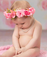 baby sitting on a pink surface with a floral headband and pearl necklace against a blurred background.